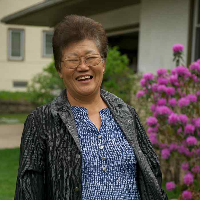 Smiling older Asian woman standing in front of a house and flowering bush