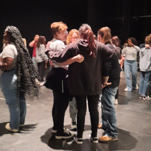 A group of high school students stand in circle, arms around one another. Other student move around the space in the background.