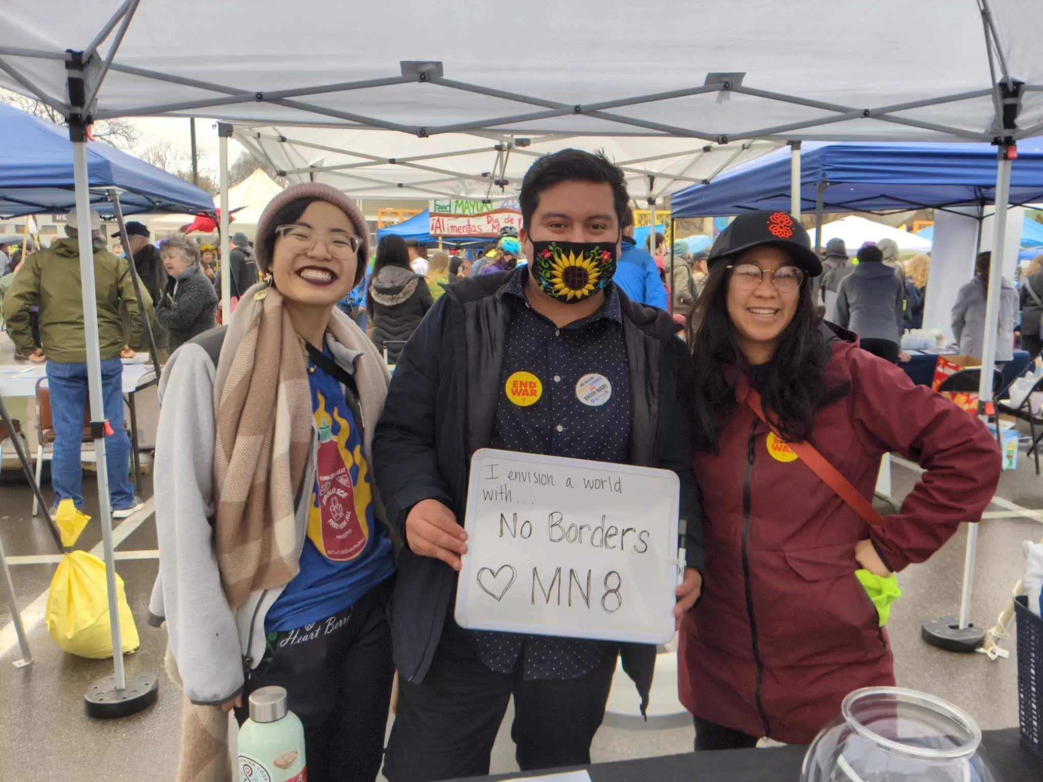 A woman, man and woman stand together with event tents behind them. The man holds a sign reading I envision a world with no borders.