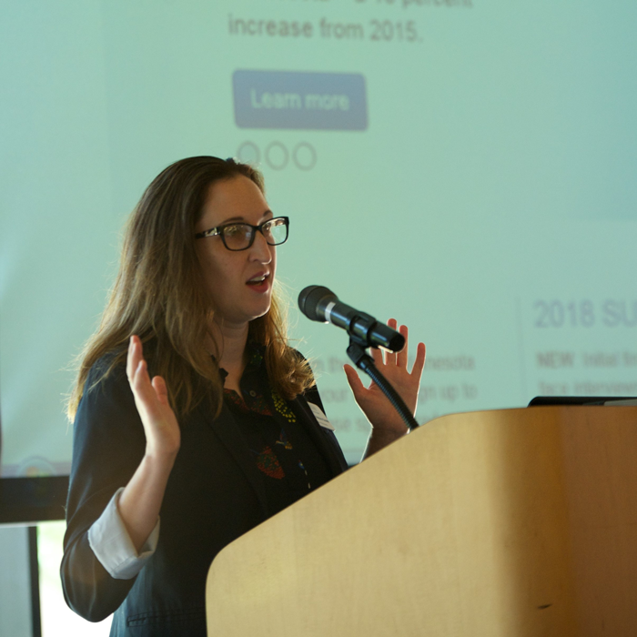 A woman speaks from a podium with a large screen behind her.