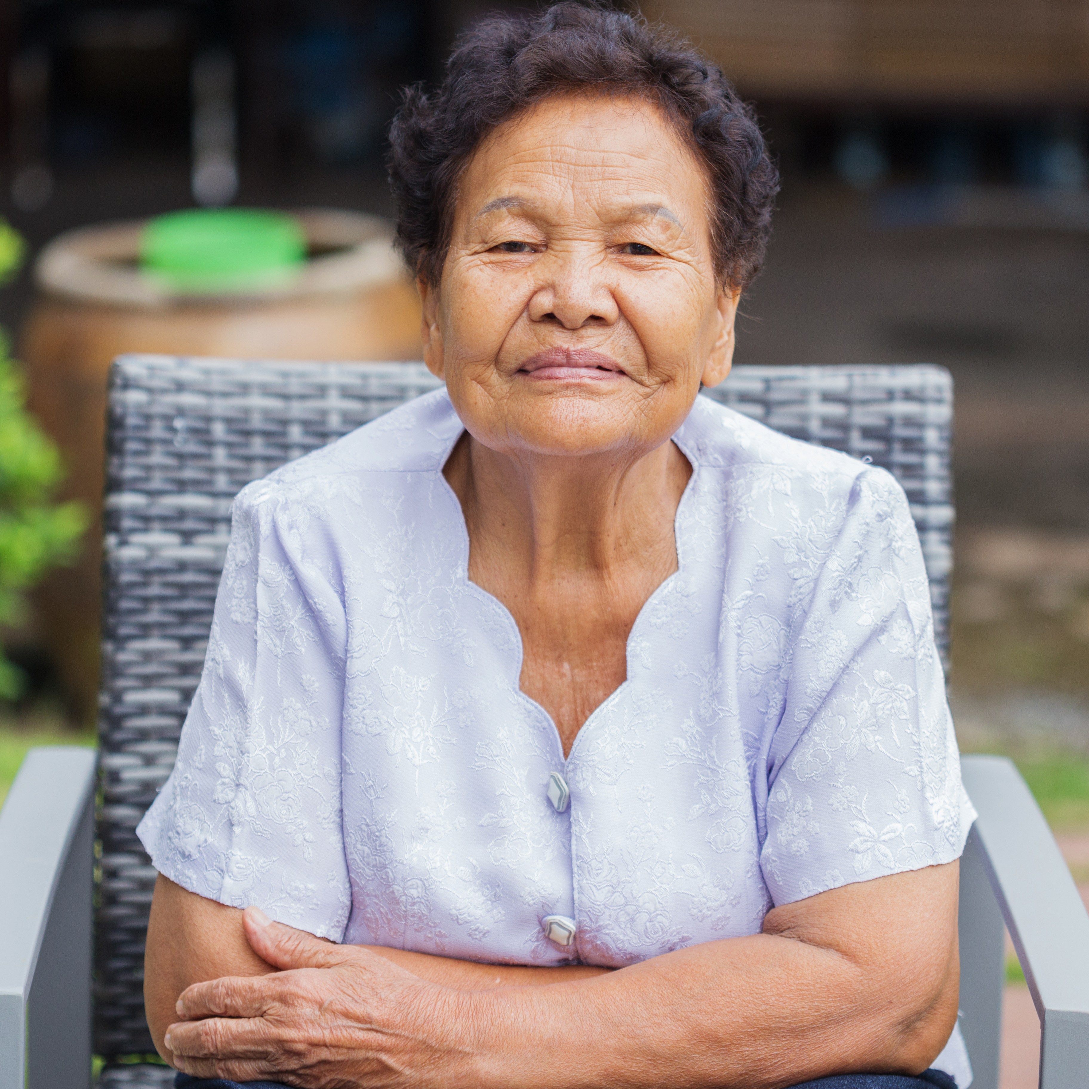 An older woman sitting in a patio chair leans forward toward the camera with arms crossed.  