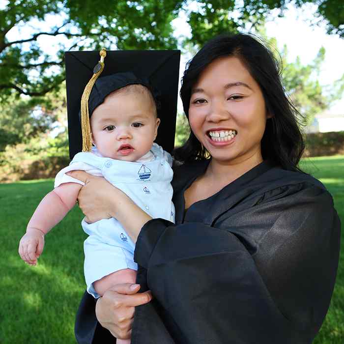 A mother in a graduation gown holds a baby wearing a graduation cap.