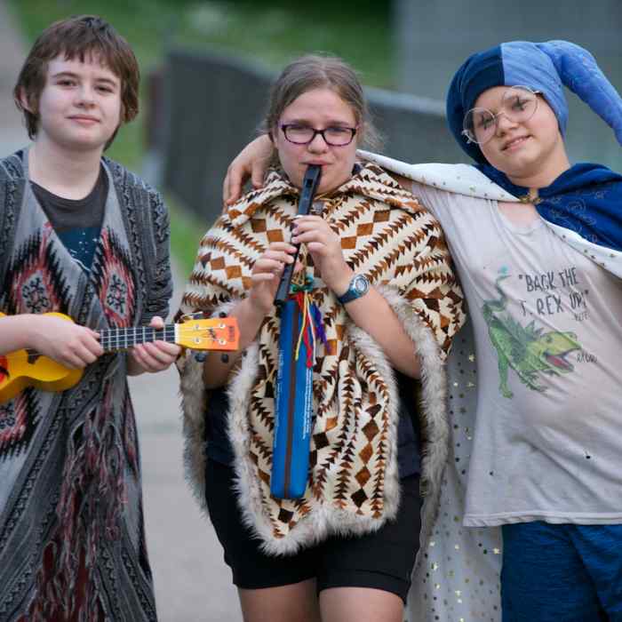 Three young people holding musical instruments stand together on a sidewalk.
