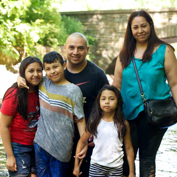 A family of two adults and three children wades in a stream in a park setting.