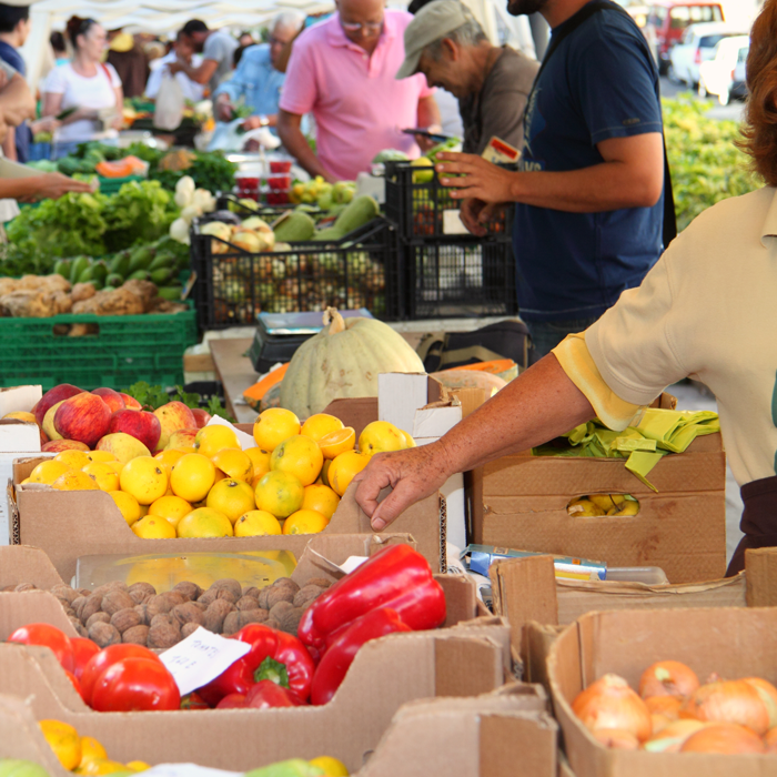 A woman reaches across boxes of produce while several people browse vegetables in the background.
