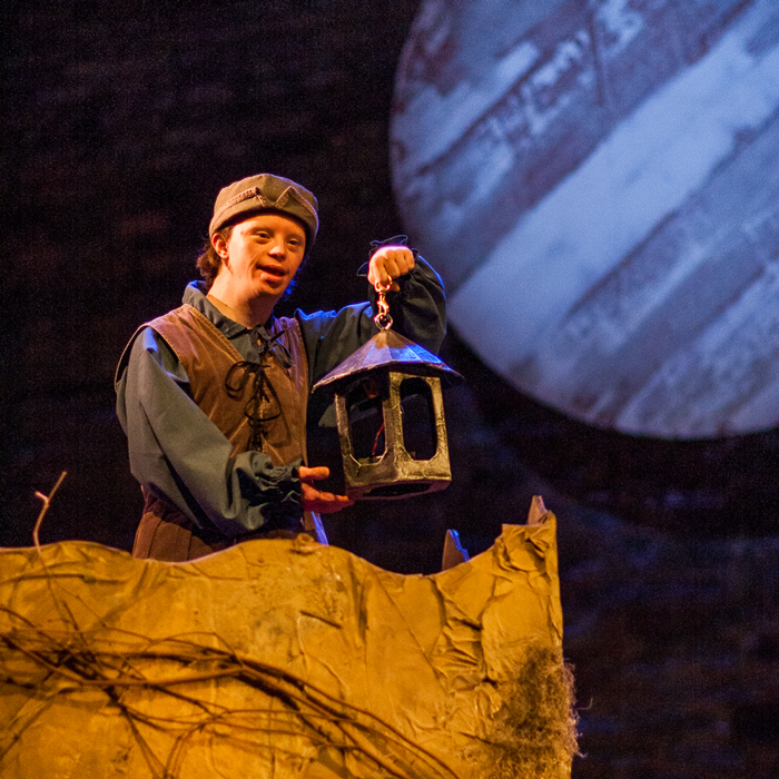 An actor holds a lantern while performing on stage, with a large moon over their shoulder.