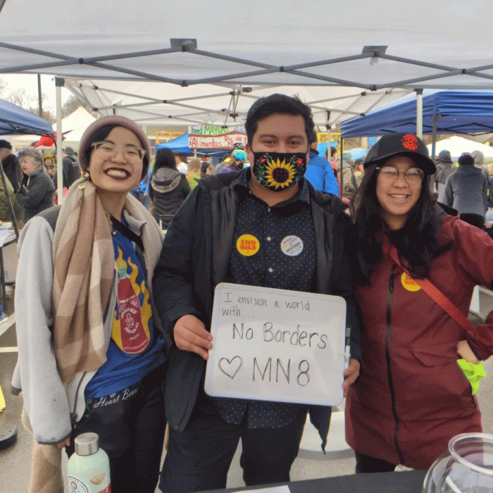 Three people stand side-by-side under a pop-up tent at an outdoor festival. The middle person holds a sign that says I envision a world with no borders -- MN8.