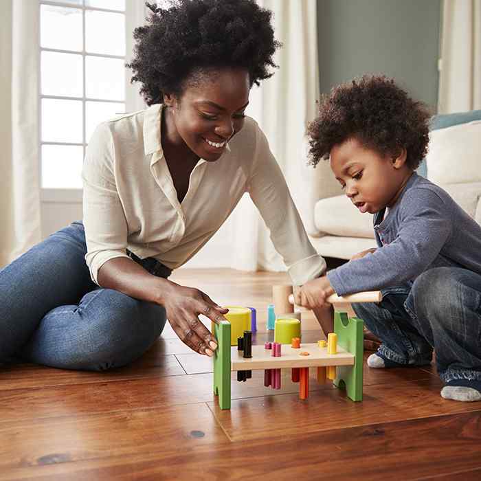 A woman and a toddler sit on the wood floor of a home and play with multi-colored toys. 