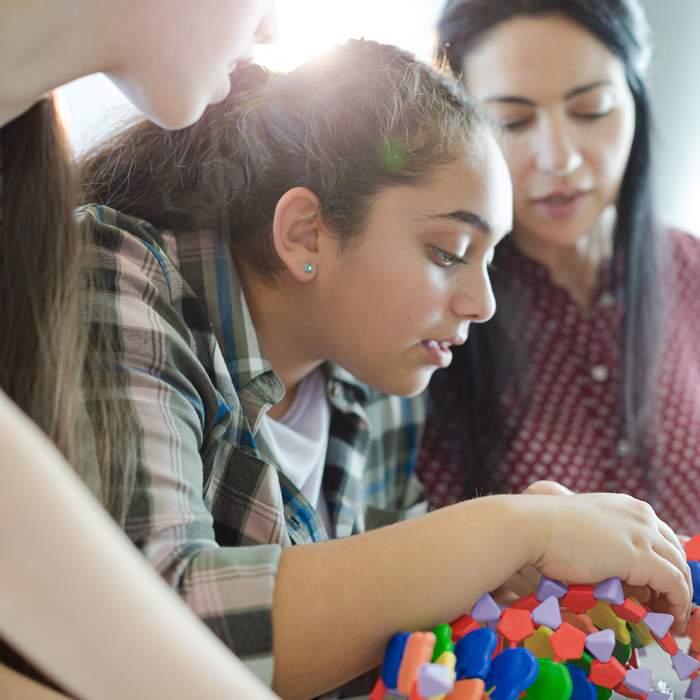 A high school student engages in a hands-on project while a teacher and fellow student look on.