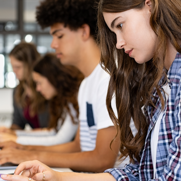 A row of high school age students work at laptops in a classroom setting.