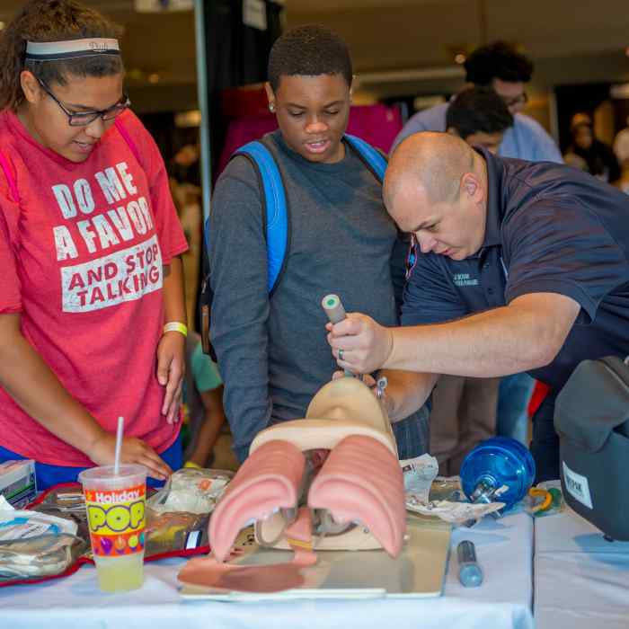 Two youth look on as a man shows them how to open the airway on a demonstration mannequin.