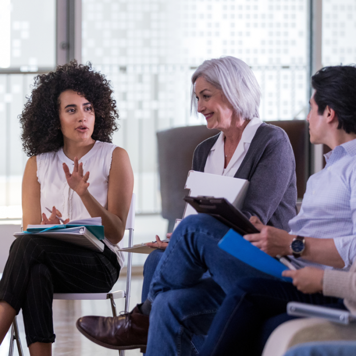 Three people sit in an office space with notebooks and pens in hand. One person is gesturing with her hand, while others listen.