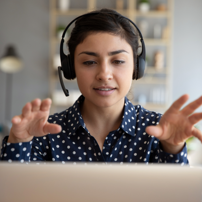 A woman wearing a headset talks as she faces a laptop. Her hands are raised as she talks.