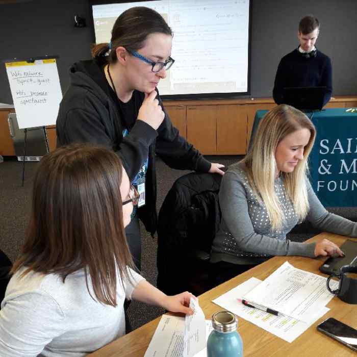 Three women huddle around a table, looking at a computer.