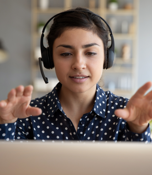 A woman wearing a headset talks as she faces a laptop. Her hands are raised as she talks.