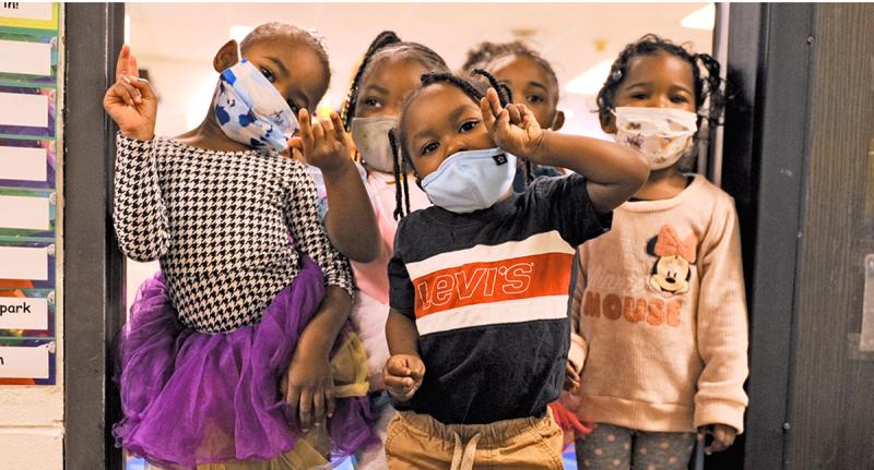 A photo of five young children standing inside a classroom doorway, looking at the camera and posing. They are wearing facemasks over their noses and mouths.