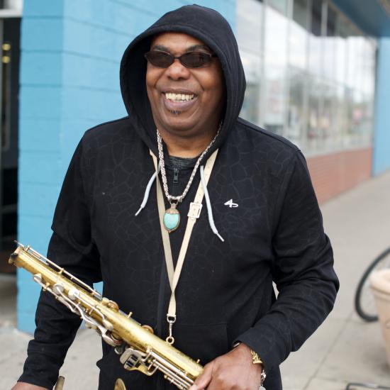 An adult stands on the sidewalk in front of a brick building, smiling and holding a saxophone.