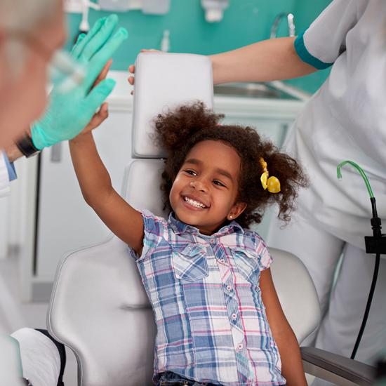 A young girl, sitting in a dentist's chair, smiles and gives a high five to a man in soft focus.