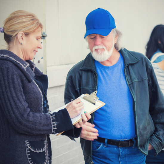 A woman holding writes on a clipboard as a man facing her responds.
