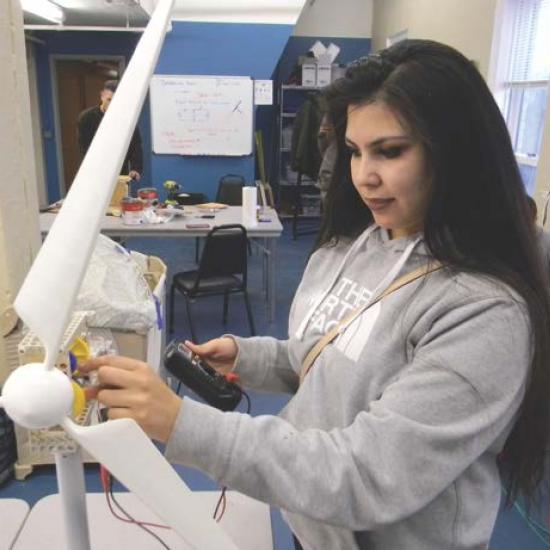 A youth checks the gears on a tabletop windmill with one hand, while she holds control board in the other.