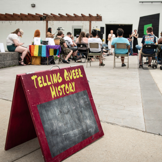 A Telling Queer History sign sits in the foreground, with a circle of event attendees gathered behind.