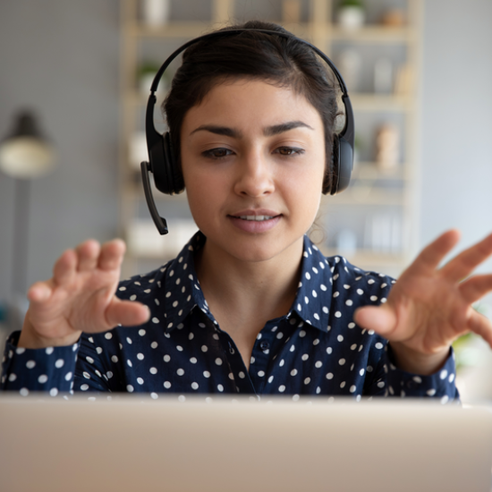 A woman wearing a headset talks as she faces a laptop. Her hands are raised as she talks.