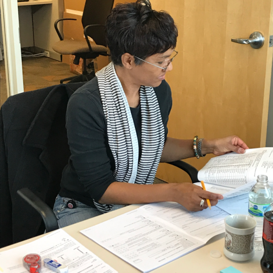 A woman is seated at a table with stacks of survey booklets in front of her. She holds a pencil as she flips through a survey. 