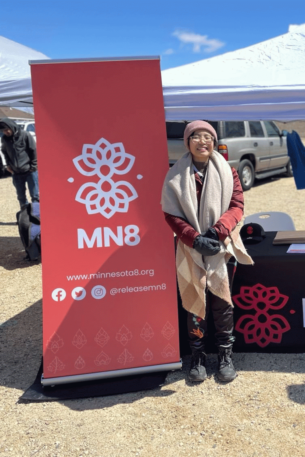 A person stands next to a red MN8 banner at an outdoor celebration.