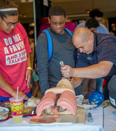Two youth look on as a man shows them how to open the airway on a demonstration mannequin.