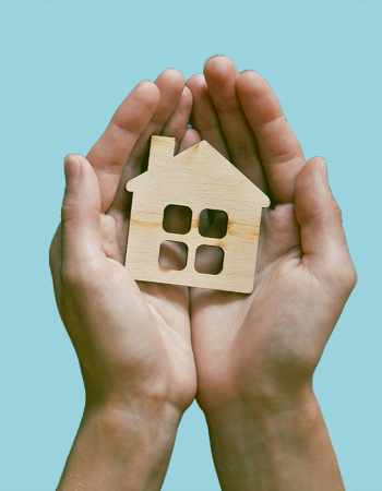 A pair of hands hold a small cutout of a wood house.