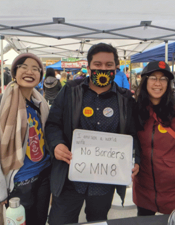 Three people stand side-by-side under a pop-up tent at an outdoor festival. The middle person holds a sign that says I envision a world with no borders -- MN8.