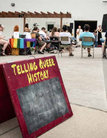 A Telling Queer History sign sits in the foreground, with a circle of event attendees gathered behind.