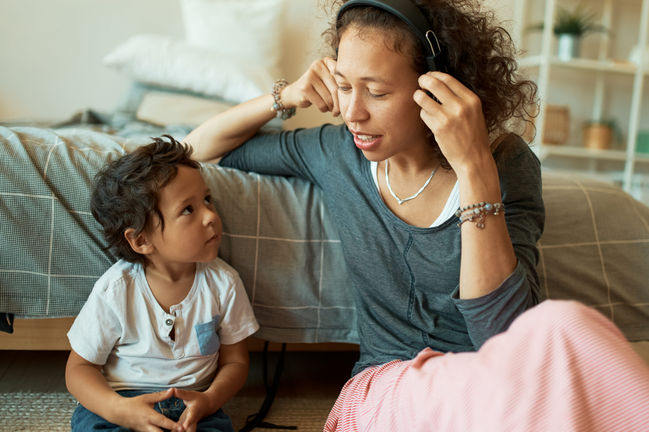 Horizontal shot of beautiful young Hispanic woman listening to music in wireless headphones sitting on floor with her handsome little son. Happy family having fun at home
