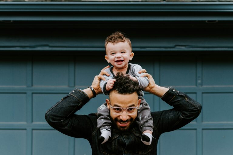 A dad holding a baby on his shoulders in front of a garage